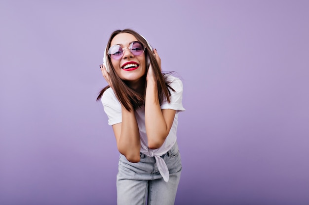 Chica alegre en gafas de sol tocando sus auriculares con una sonrisa. Foto interior de hermosa modelo femenina en jeans aislado.