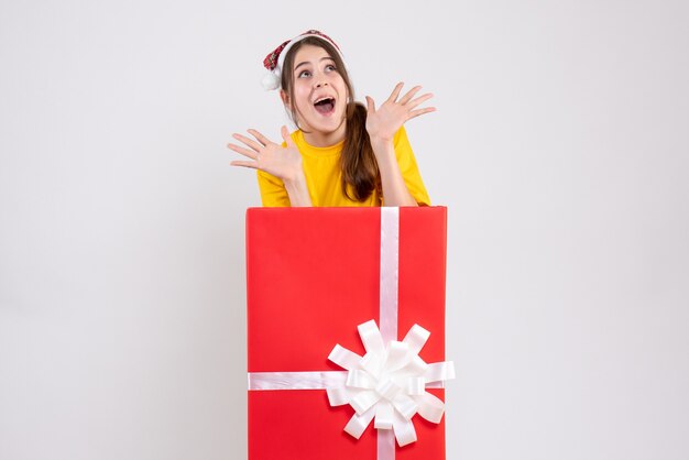 Chica alegre con gorro de Papá Noel de pie detrás de un gran regalo de Navidad en blanco