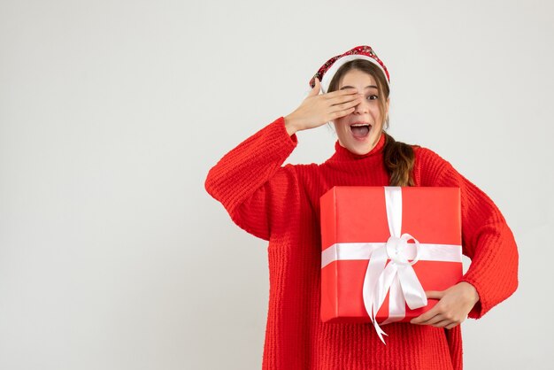 Chica alegre con gorro de Papá Noel que cubre sus ojos con la mano en blanco