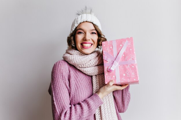 Chica alegre en gorro de punto y bufanda con caja rosa con cinta. Feliz joven mujer de pelo corto con regalo de año nuevo posando con una sonrisa en la pared blanca.