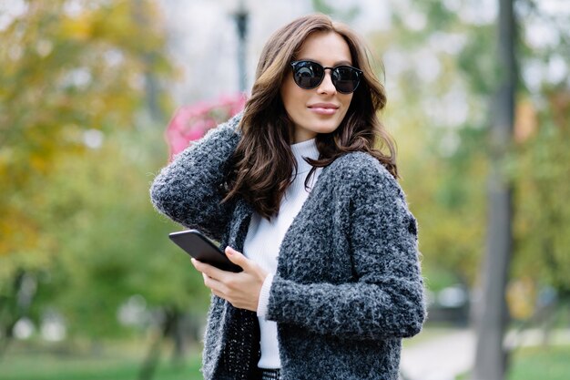 Chica alegre guapa con cabello corto y oscuro en elegante jersey gris disfrutando el fin de semana, pasando tiempo al aire libre. Atractiva mujer joven con peinado de moda caminando por la calle por el parque