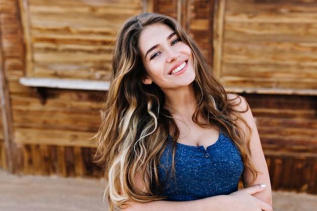 Chica alegre guapa con cabello rizado con elegante atuendo azul oscuro disfrutando el fin de semana, pasando tiempo al aire libre. Mujer joven atractiva con peinado de moda caminando por la calle por puesto de madera