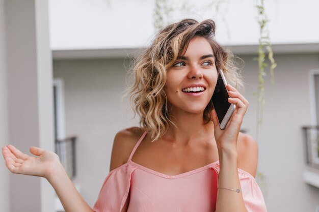 Chica alegre hablando por teléfono y soñadora mirando a otro lado. Retrato de primer plano de una dama europea bronceada en blusa rosa llamando a alguien mientras está de pie en el balcón del hotel.