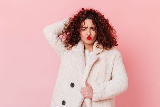 Chica alegre con labios rojos toca su cabello y hace una expresión facial divertida. Mujer en bata blanca posando en espacio rosa.