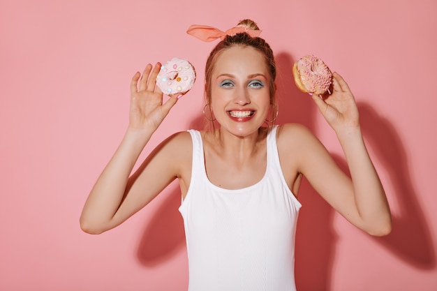 Foto gratuita chica alegre con peinado rubio en aretes y maquillaje fresco en ropa blanca sonriendo y sosteniendo dos donas en la pared aislada