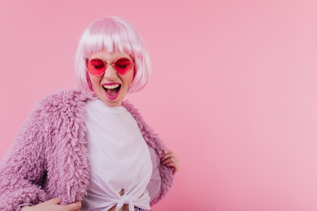 Chica alegre en peluca bailando en la pared rosa. Mujer joven emocionada en peruke expresando felicidad