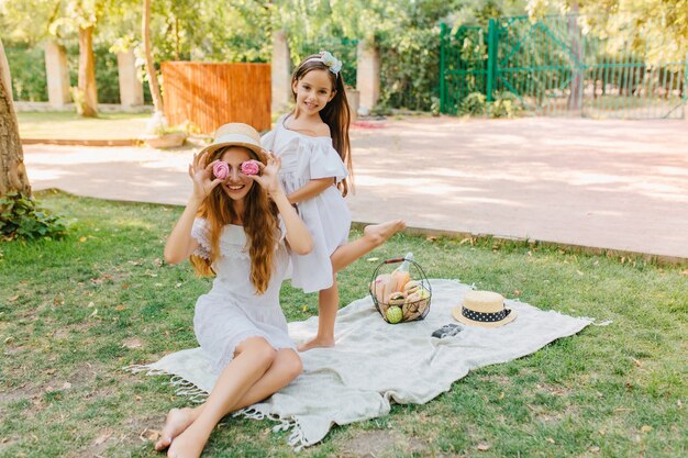 Chica alegre de pie sobre una pierna mientras su madre divertida jugando con galletas. Retrato al aire libre de broma mujer de pelo largo disfrutando de un picnic con su hija en vacaciones.