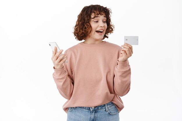 Chica alegre y rizada, se ve emocionada con la tarjeta de crédito, sostiene el teléfono inteligente en la mano, hace un pedido en línea, compra en una tienda de Internet, está feliz contra el fondo blanco