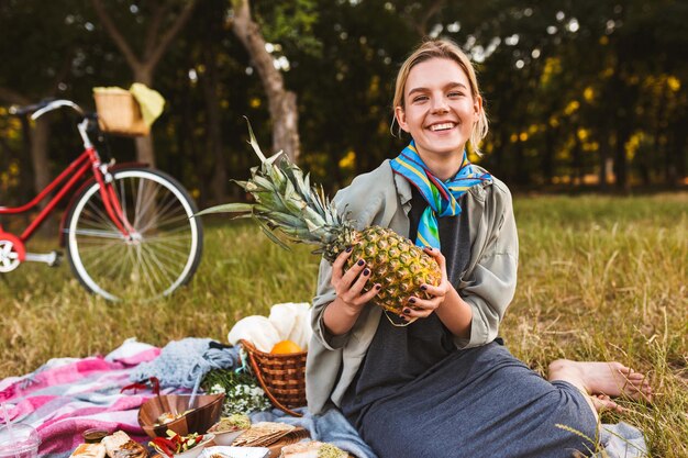 Chica alegre sentada en una manta de picnic en el césped felizmente sosteniendo piña en las manos pasando tiempo en el parque