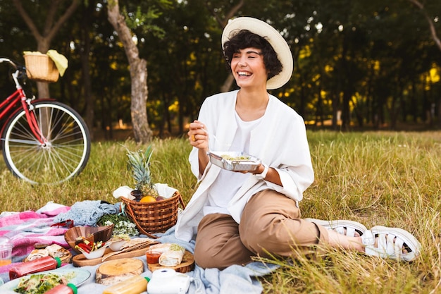 Chica alegre sentada con sombrero y camisa blanca con ensalada en las manos felizmente riendo pasando tiempo en un picnic en el parque