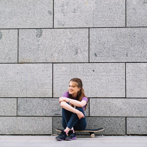 Chica alegre se sienta en una patineta en frente de una pared de ladrillos