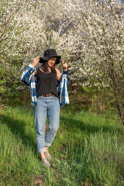 Chica alegre con un sombrero entre los árboles en flor en la primavera, en un estilo casual.