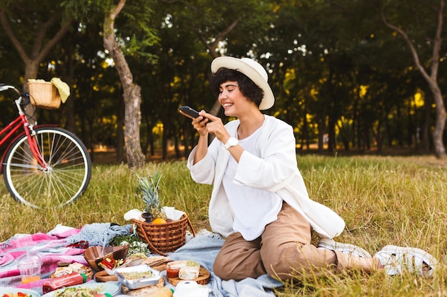 Foto gratuita chica alegre con sombrero y camisa blanca felizmente tomando fotos en el celular de comida de picnic pasando tiempo en el parque