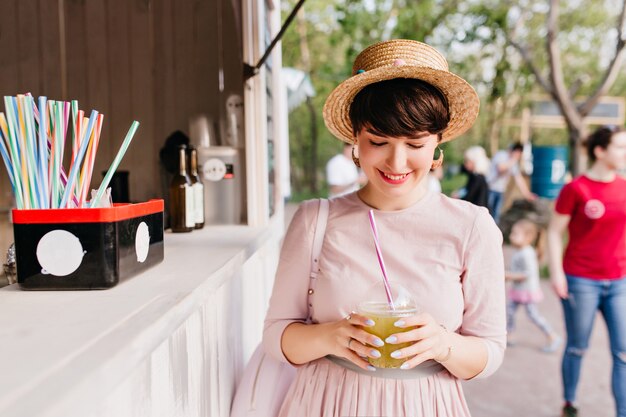 Chica alegre con sombrero de paja compró una bebida fría, caminando por la plaza de la ciudad