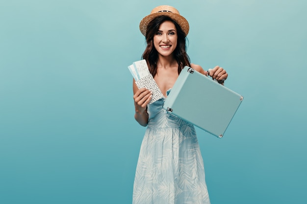 Chica alegre con sombrero de paja sosteniendo boletos y maleta sobre fondo azul.