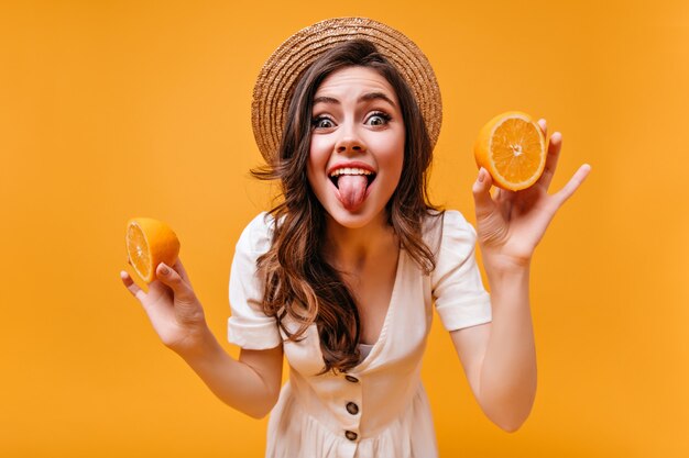 Chica alegre con sombrero y vestido de verano muestra lengua y posa con naranjas.