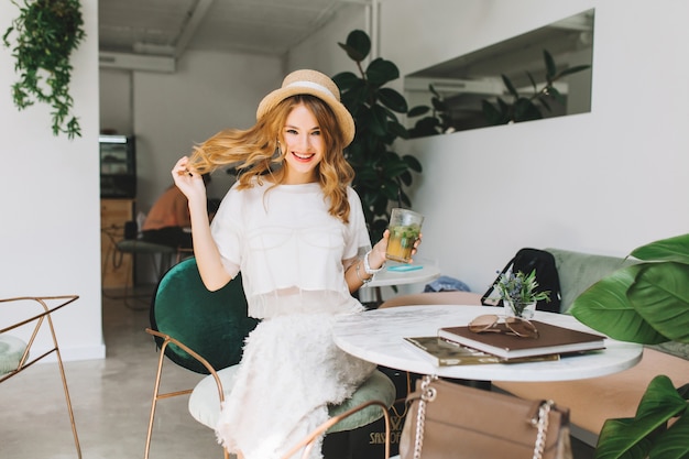 Foto gratuita chica alegre en sombrero vintage de moda jugando con cabello rubio y riendo relajándose en el café