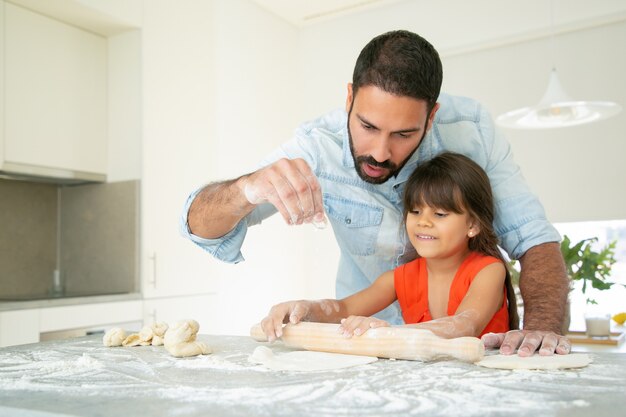 Chica alegre y su papá amasando y rodando masa en la mesa de la cocina con harina desordenada.