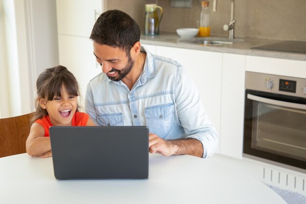 Chica alegre y su papá usando laptop para videollamadas, sentados a la mesa, viendo películas divertidas, mirando la pantalla.