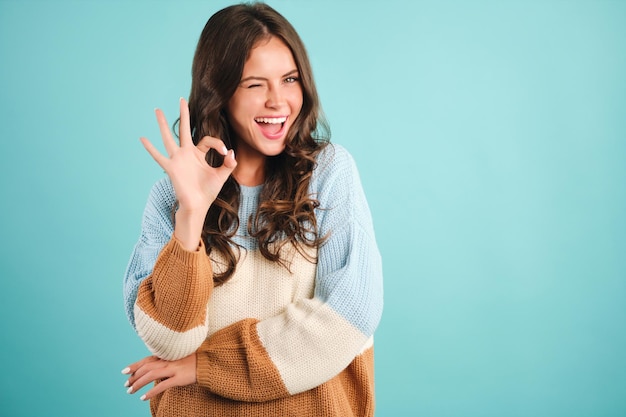 Una chica alegre con un suéter acogedor mostrando felizmente un gesto correcto y guiñando un ojo en la cámara sobre el fondo azul.