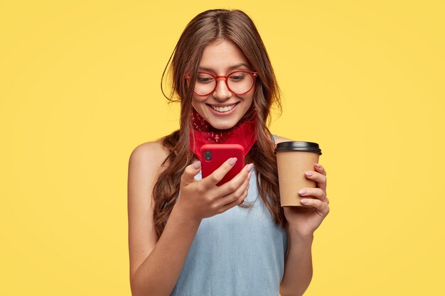 Chica alegre tiene una pausa para el café, se regocija comprando un nuevo dispositivo, lee la notificación en el teléfono móvil rojo, actualiza la aplicación favorita, escribe mensajes y sonríe en la pantalla, usa gafas, aislado sobre una pared amarilla