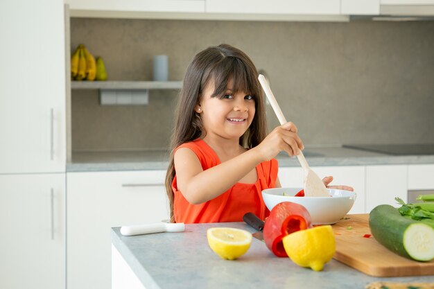 Chica alegre tirando ensalada en un tazón con cuchara de madera grande. Niño lindo pasar tiempo en casa durante la pandemia, cocinar verduras, posando, sonriendo a la cámara. Aprender a cocinar concepto