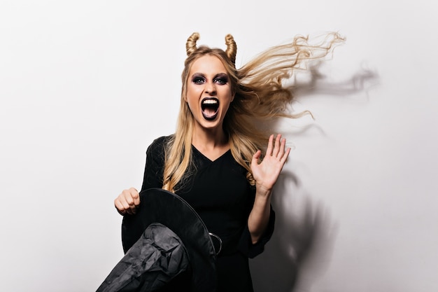 Foto gratuita chica alegre en traje de carnaval posando en la pared blanca. bruja malvada divirtiéndose en halloween.