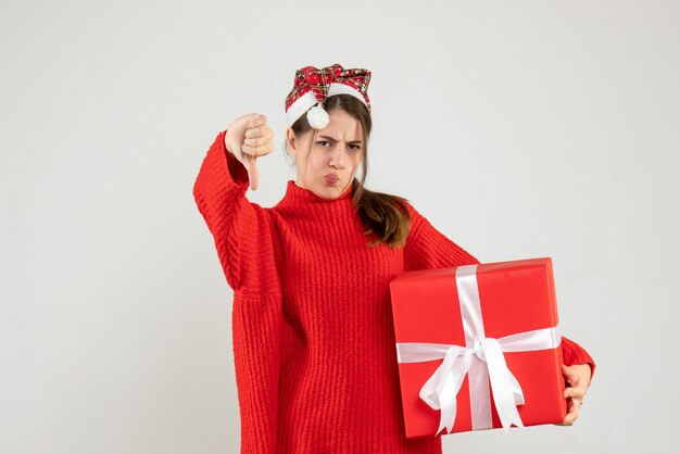 Chica sin alegría con gorro de Papá Noel sosteniendo presente haciendo pulgar hacia abajo firmar de pie en blanco