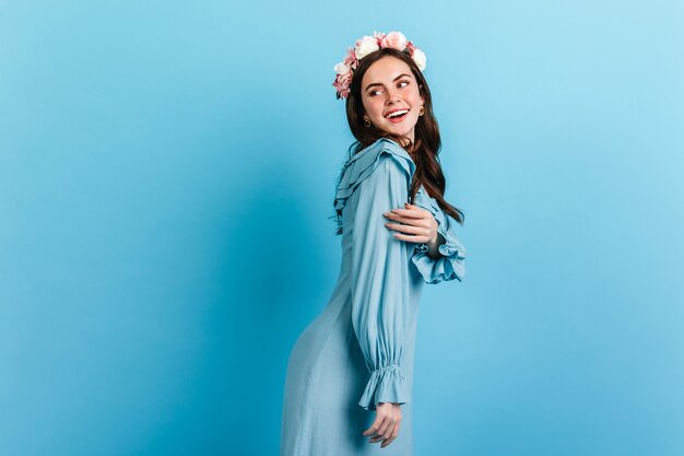 Una chica amable con una sonrisa blanca como la nieve mira a su alrededor. Modelo con corona de flores y vestido de seda posando en pared azul.