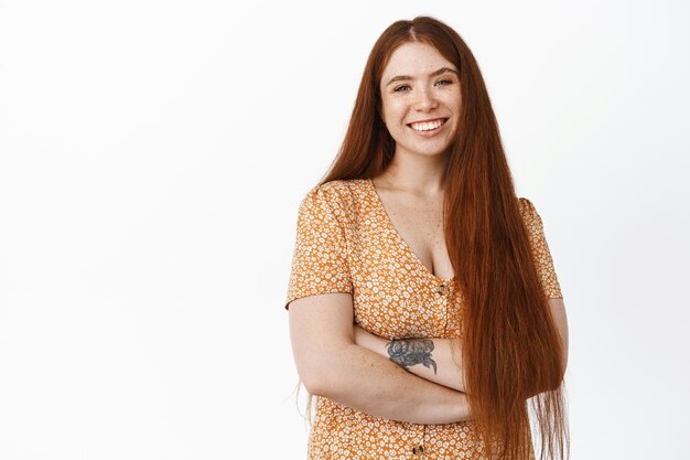 Chica ambiciosa feliz con el pelo largo y rojo sonriendo y riendo cruzando los brazos sobre el pecho posando vestida sobre fondo blanco