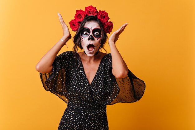 Chica de América Latina con maquillaje de calavera reacciona emocionalmente y posa para retrato en pared naranja