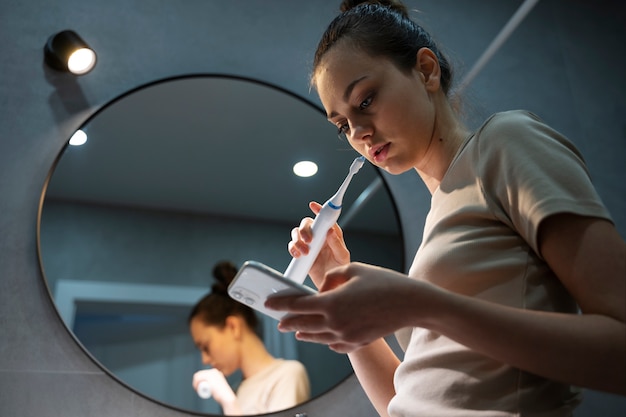 Chica de ángulo bajo cepillando dientes con teléfono inteligente