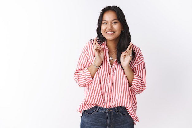 Chica anticipando con entusiasmo por el día de San Valentín cruza los dedos para la buena suerte, la fecha de la esperanza va bien sonriendo emocionada posando esperanzada, orando por hacerse realidad contra la pared blanca