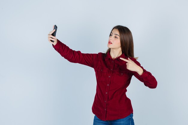 chica apuntando a la pantalla del teléfono inteligente mientras toma selfie con camisa burdeos y parece confiada.