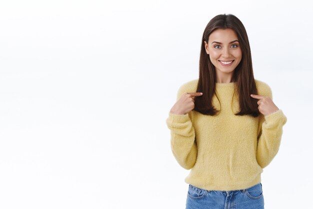 Chica apuntando a sí misma y sonriendo como sugerencia de ayuda propia, candidatura personal, expresando su voluntad de participar en el evento, sonriendo y luciendo lista para las tareas, de pie en la pared blanca