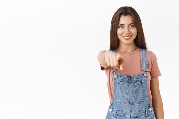 Chica apuntando hacia ti como haciendo una elección, reúne al equipo. Atractiva cámara indicadora femenina linda confiada y motivada con expresión satisfecha y segura de sí misma, fondo blanco de pie