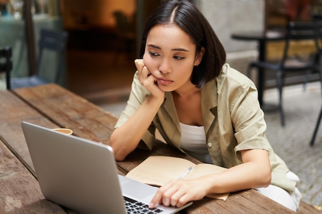 Chica asiática aburrida sentada en un café y mirando la computadora portátil estudiando y sintiéndose triste haciendo la tarea