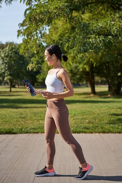 Chica asiática activa en entrenamiento de ropa de fitness en el parque caminando en ropa deportiva con teléfono inteligente y agua
