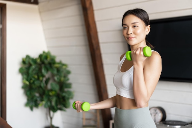 Chica asiática activa y saludable con cuerpo en forma haciendo ejercicios de fitness en casa levantando pesas ...