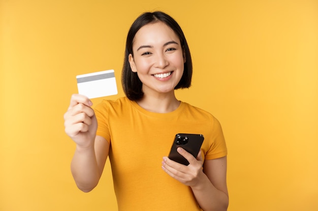 Chica asiática alegre sonriendo mostrando tarjeta de crédito y teléfono inteligente recomendando la banca móvil de pie contra el fondo amarillo