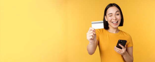 Chica asiática alegre sonriendo mostrando tarjeta de crédito y teléfono inteligente recomendando la banca móvil de pie contra el fondo amarillo