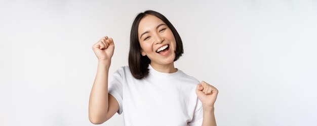 Chica asiática bailando celebrando sentirse feliz y optimista sonriendo ampliamente de pie sobre el fondo blanco del estudio