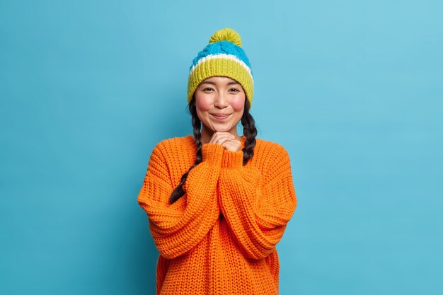 La chica asiática bonita mantiene las manos debajo de la barbilla se pone roja y tímida mientras recibe el cumplido de su novio viste un suéter de punto cálido y un sombrero tiene una mirada encantadora aislada sobre una pared azul