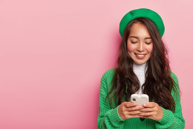 Chica asiática bonita de moda sostiene el teléfono móvil, vestida con ropa verde, navega por internet en un teléfono celular moderno, envía un mensaje de texto