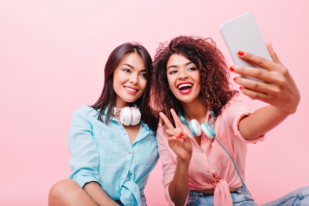 Chica asiática bronceada complacida sonriendo suavemente mientras su amiga africana hace selfie. Retrato interior de mujer negra alegre con smartphone tomando una foto de sí misma cerca de la dama hispana.