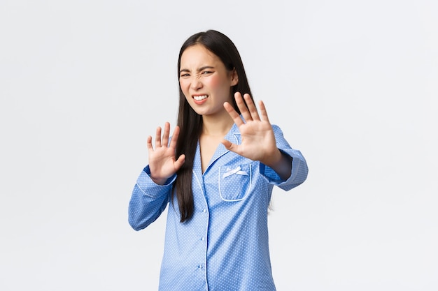 Chica asiática cavada y disgustada en pijama azul extiende las manos hacia adelante en señal de rechazo, rechazando algo repugnante, haciendo muecas de aversión y disgusto, dándose la mano en negación sobre fondo blanco.