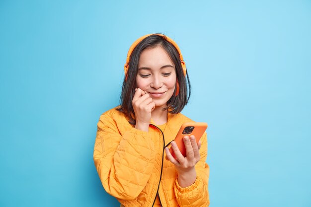 Chica asiática complacida con cabello oscuro enfocado en la pantalla del teléfono inteligente elige la pista de audio de la lista de reproducción usa auriculares estéreo inalámbricos en las orejas vestidas con una chaqueta naranja aislada sobre una pared azul