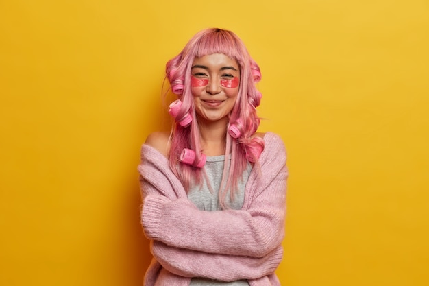 Chica asiática complacida se siente cómoda con un jersey de lana, se abraza y sonríe suavemente, tiene el cabello rosado, se peina con rulos, se aplica almohadillas de belleza