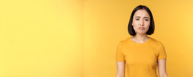 Una chica asiática decepcionada que se enfurruña y se ve molesta se siente sin aire de pie en una camiseta amarilla sobre un fondo blanco