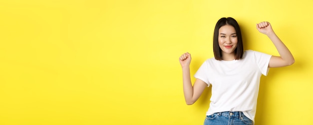 Chica asiática despreocupada bailando y divirtiéndose posando en camiseta blanca sobre fondo amarillo
